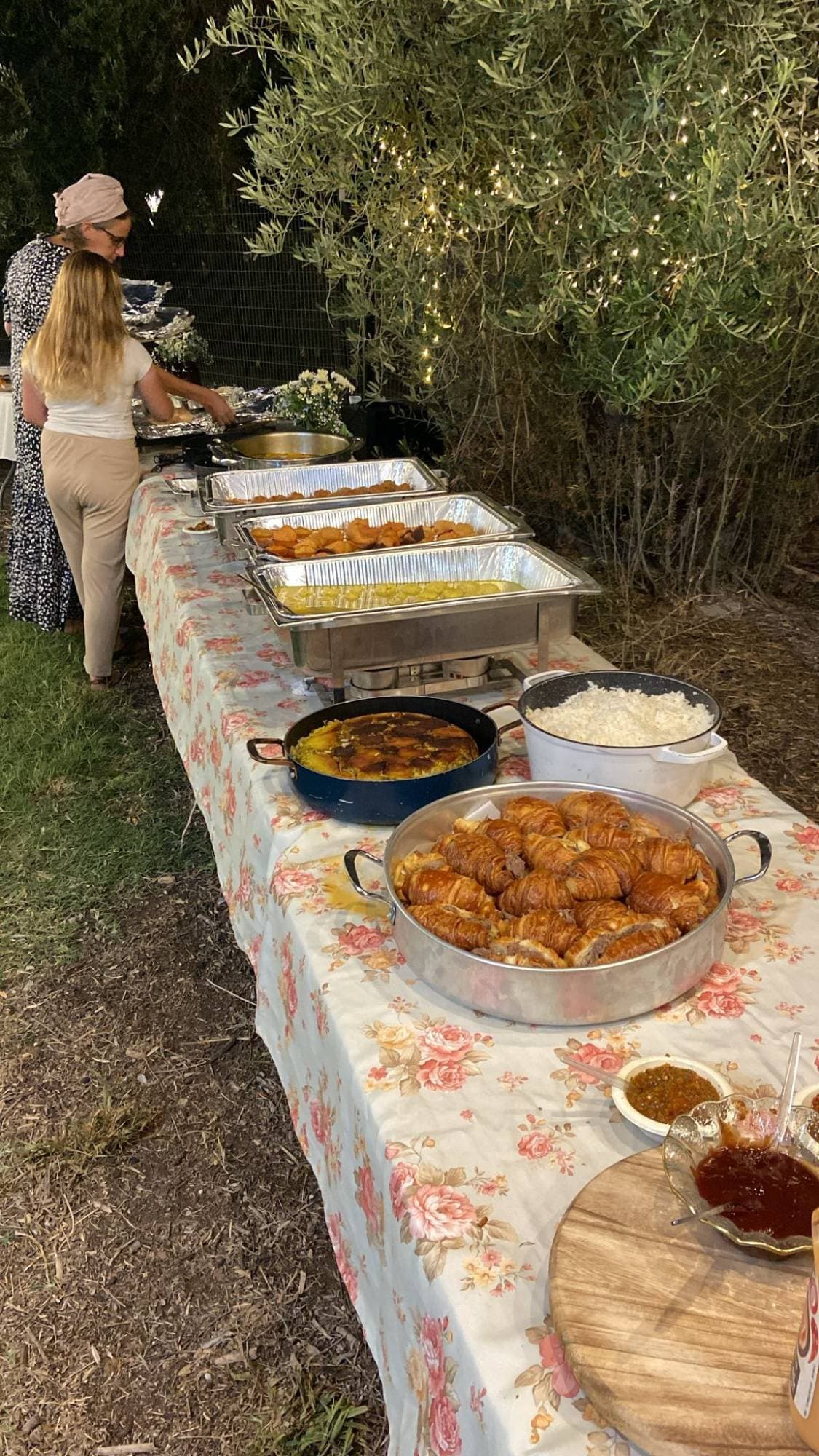 ערב קייטרינג של בישולילים בגן — שולחן ארוך עם מגשי הגשה חמים, אורז, קרואסונים וסלטים תחת עצי זית ואורות