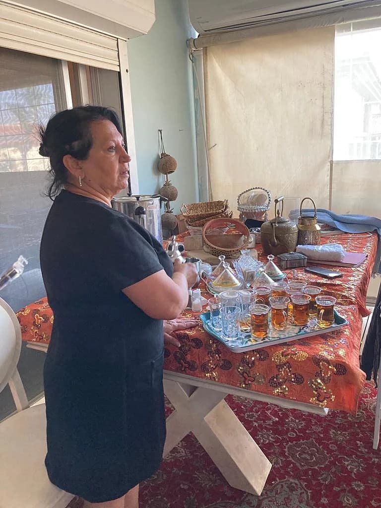 לילי טויזר עומדת ליד שולחן תה פרסי מסורתי עם קומקום נחושת מגולף ומגש כוסות תה