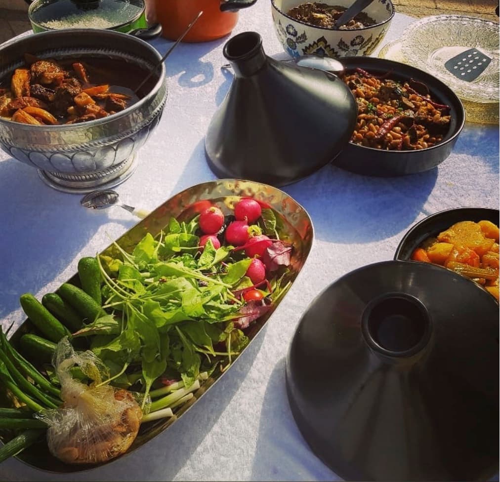 שולחן חוץ של בישולילים בשמש — מגש סלט ירקות ונוביה, קערה מגולפת של תבשיל פירות יבשים עם בשר, טאג׳ין שחור פתוח עם חיטה ותבלינים, וקדרת ירקות שורש מבושלת