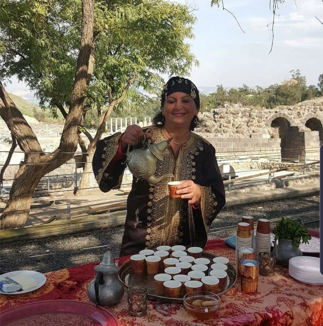 לילי טויזר בתלבושת פרסית מסורתית מוזגת תה על רקע עתיקות בית שאן
