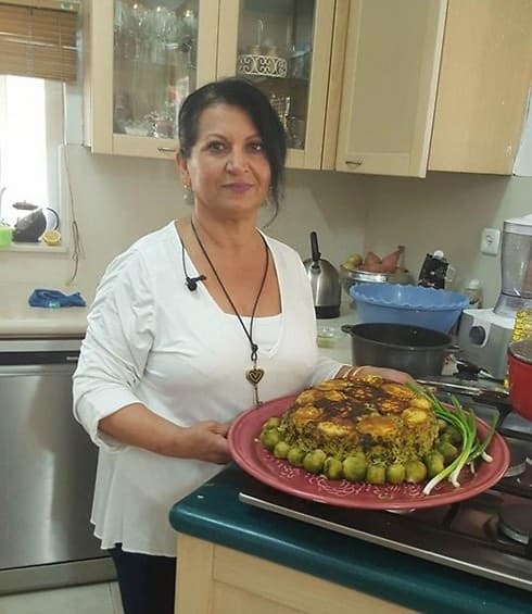 לילי טויזר מחייכת במטבחה, אוחזת מגש אדום עם אורז פרסי, אפונה ירוקה ובצל ירוק